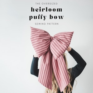 May include: A large, striped, pink bow sewing pattern. The bow is held up in front of a person. The text on the image reads "THE OVERSIZED heirloom puffy bow SEWING PATTERN."
