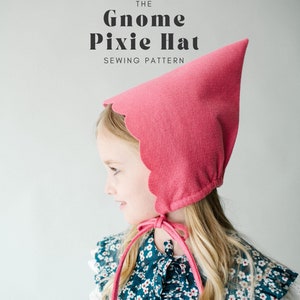 Può includere: Un cappello da gnomo pixie in feltro rosa con un bordo a scalare e una cravatta. Il cappello è indossato da un bambino con lunghi capelli biondi e un colletto floreale blu e bianco.