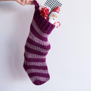 May include: A handmade, striped purple and lavender knitted Christmas stocking filled with gifts. The stocking is held by a hand and contains a wrapped gift, a Santa-shaped chocolate, a red bow, and a candy cane. Text at the top reads "Easiest Ever Stocking Knitting Pattern."
