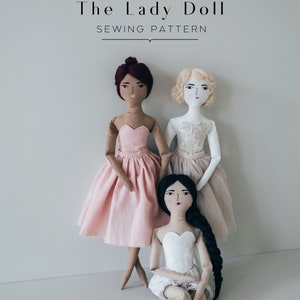 Peut inclure: Trois poupées en tissu faites à la main avec des couleurs de cheveux et des tenues différentes. Les poupées portent des robes et ont des visages peints. Les poupées sont debout et assises sur un fond blanc. Le texte "The Lady Doll SEWING PATTERN" est en haut de l'image.