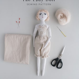 May include: A sewing pattern for a handmade doll, "The Lady Doll". The image shows a doll with blonde hair, a white lace bodice, and beige pants. The doll is surrounded by sewing supplies, including fabric, scissors, needles, and stuffing.