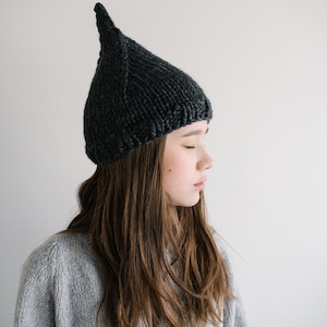 May include: A person wearing a dark gray knitted hat with a pointed top. The hat is made of a thick yarn and has a simple design. The person is wearing a gray sweater.