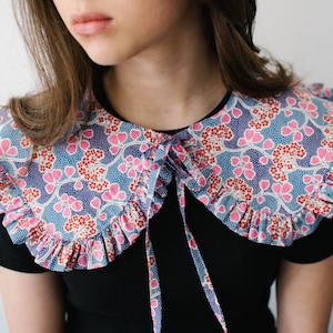 K&ouml;nnte beinhalten: Ein gro&szlig;er, gekr&auml;uselter Kragen mit einem blauen und rosa Blumendesign. Der Kragen ist vorne mit einem schwarzen Band gebunden.