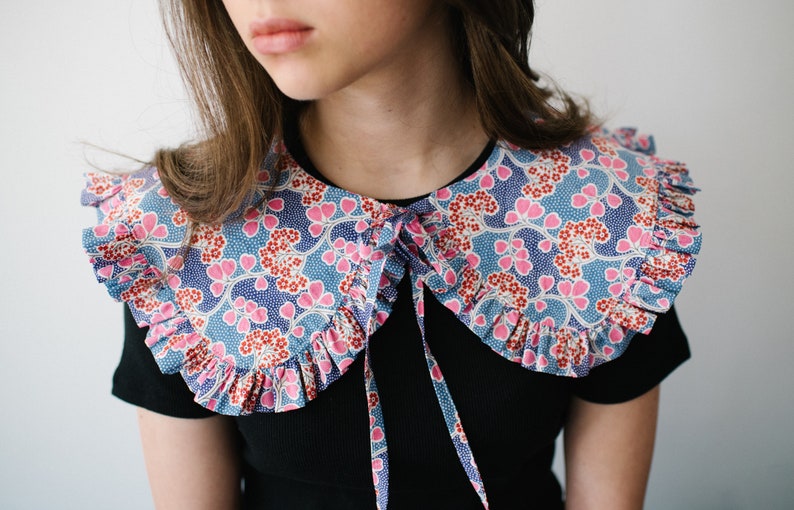 K&ouml;nnte beinhalten: Ein gro&szlig;er, gekr&auml;uselter Kragen mit einem blauen und rosa Blumendesign. Der Kragen ist vorne mit einem schwarzen Band gebunden.