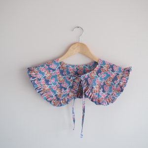 K&ouml;nnte beinhalten: Ein blauer und rosa floral gemusterter Stoffkragen mit R&uuml;schen und einer Schleifenbindung.