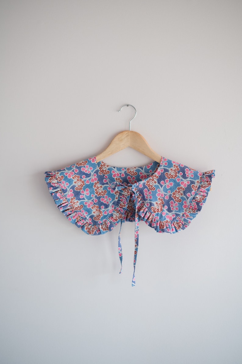 K&ouml;nnte beinhalten: Ein blauer und rosa floral gemusterter Stoffkragen mit R&uuml;schen und einer Schleifenbindung.