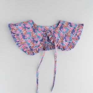 K&ouml;nnte beinhalten: Ein blauer und rosa floral bedruckter Stoffkragen mit gekr&auml;uselten R&auml;ndern und zwei langen B&auml;ndern.