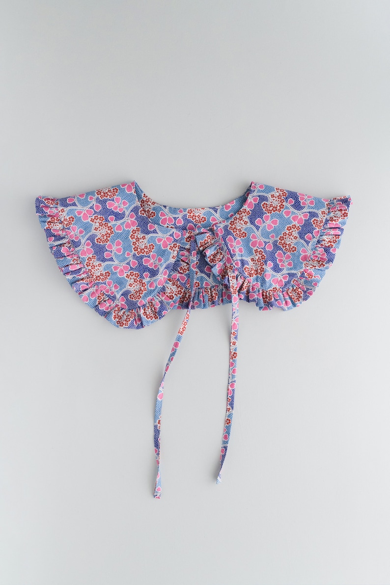 K&ouml;nnte beinhalten: Ein blauer und rosa floral bedruckter Stoffkragen mit gekr&auml;uselten R&auml;ndern und zwei langen B&auml;ndern.