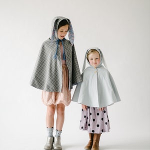 May include: Two children wearing bunny-themed capes. The older child is wearing a grey plaid cape with floral bunny ears and a pink dress. The younger child is wearing a light blue cape with white bunny ears and a purple polka dot skirt.