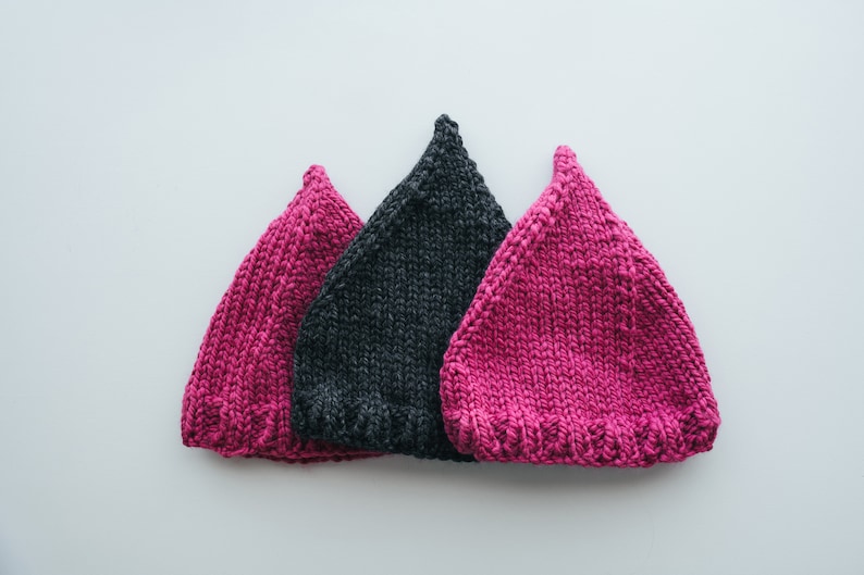 May include: Three knitted hats, one dark gray and two pink, arranged in a triangle shape on a white background.