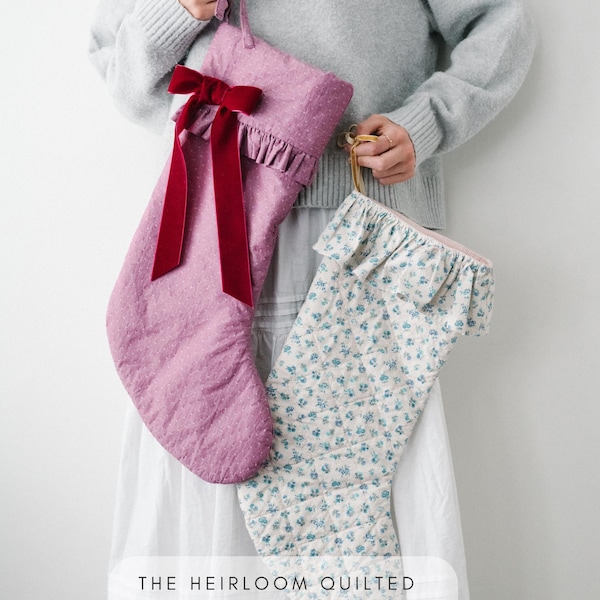May include: Two quilted stockings featuring a ruffled top edge. One stocking is solid mauve with a deep red velvet ribbon bow. The other is white with a small blue floral print. Text at the bottom reads: THE HEIRLOOM QUILTED stocking SEWING PATTERN.