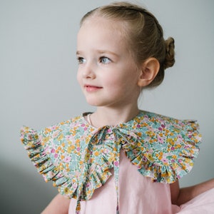K&ouml;nnte beinhalten: Ein junges M&auml;dchen tr&auml;gt ein rosa Kleid mit einem floralen Kragen. Der Kragen hat einen gekr&auml;uselten Rand und wird am Hals gebunden.