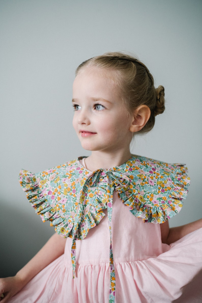 K&ouml;nnte beinhalten: Ein junges M&auml;dchen tr&auml;gt ein rosa Kleid mit einem floralen Kragen. Der Kragen hat einen gekr&auml;uselten Rand und wird am Hals gebunden.