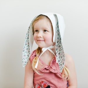 Può includere: Una bambina che indossa un cappello da coniglio bianco con fodera in tessuto floreale sulle orecchie. Il cappello ha un nastro bianco legato sotto il mento. La bambina indossa una maglietta rosa senza maniche.