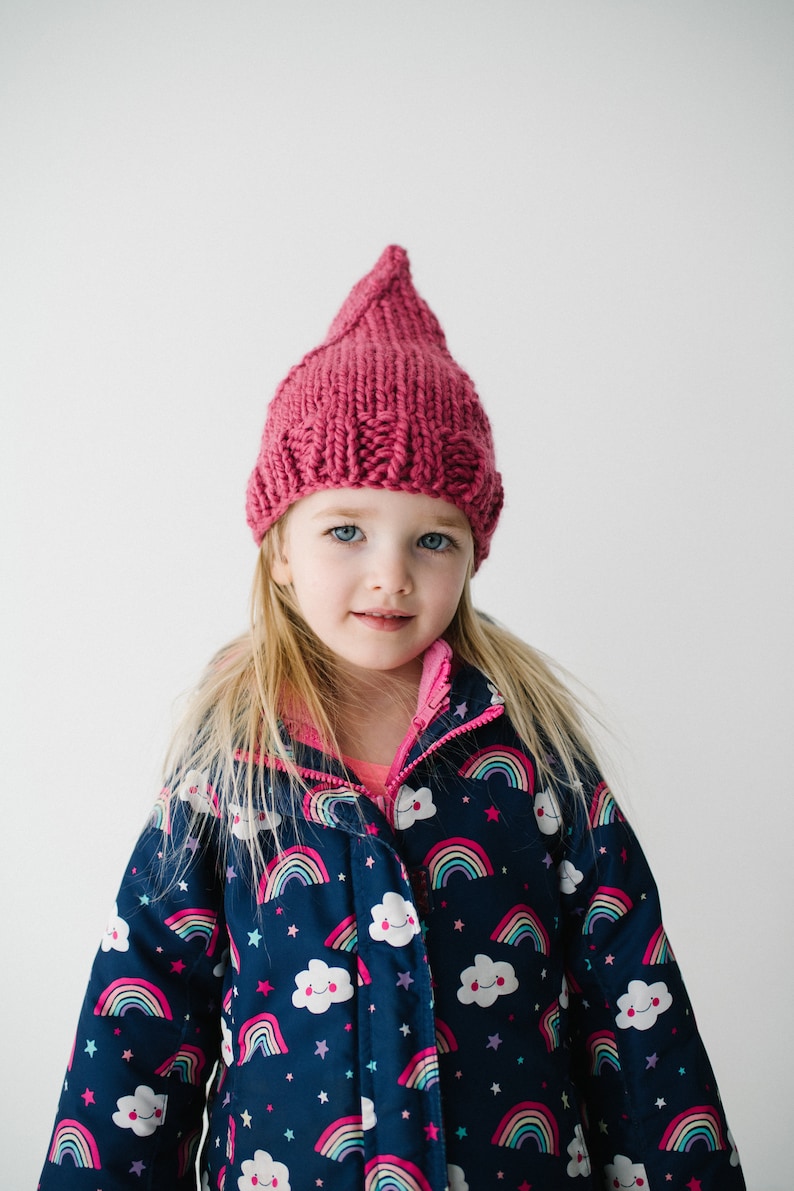 May include: A young girl wearing a pink knitted hat and a blue jacket with a rainbow and cloud pattern.