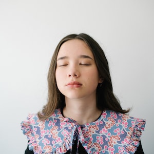 K&ouml;nnte beinhalten: Eine junge Person mit langen braunen Haaren tr&auml;gt ein schwarzes Shirt mit einem gro&szlig;en, gekr&auml;uselten, floralen Kragen. Der Kragen ist blau, rosa und wei&szlig; mit einem Blumenmuster.