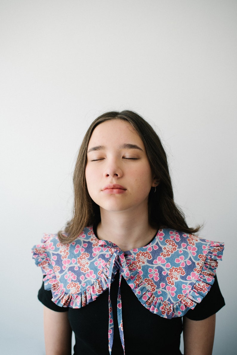 K&ouml;nnte beinhalten: Eine junge Person mit langen braunen Haaren tr&auml;gt ein schwarzes Shirt mit einem gro&szlig;en, gekr&auml;uselten, floralen Kragen. Der Kragen ist blau, rosa und wei&szlig; mit einem Blumenmuster.