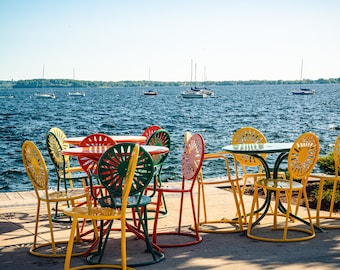 May include: A set of colorful metal chairs and tables with a view of a lake. The chairs are yellow, green, red, and orange. The tables are round and have a green top. The lake is blue and there are boats in the distance.