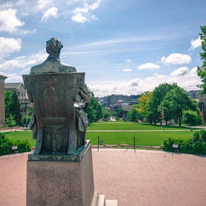 May include: A bronze statue of a man sitting on a bench, facing away from the camera. The statue is on a stone pedestal in front of a grassy lawn. There are buildings in the background, and a red flag with a white "W" on it is visible on the left side of the image.