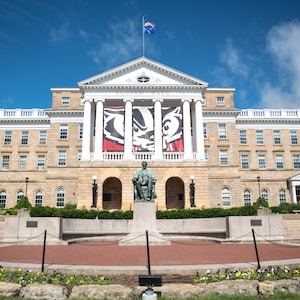 May include: A large, tan stone building with white columns and a red and black banner with a badger mascot. The building has a statue in front of it and a flagpole with an American flag.