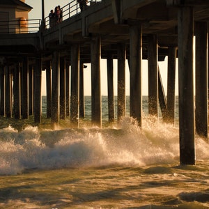 May include: A wooden pier extends over a body of water with large waves crashing against the pillars. The sun is setting, casting a golden glow on the water.
