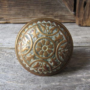 May include: A round brass door knob featuring an embossed floral pattern with a central flower motif surrounded by intricate leaf and petal designs. The metal surface displays a weathered patina with dark green oxidation in the recessed areas.
