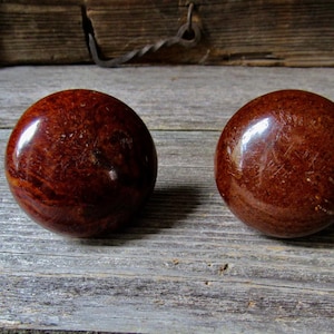 May include: Two spherical door knobs with a polished, deep reddish-brown finish. The surface of each knob displays a mottled, wood-grain-like texture with variations in color intensity, creating a glossy appearance across the rounded forms.