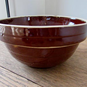 May include: A brown ceramic mixing bowl with a white rim. The bowl has a glossy finish and horizontal ridges around the exterior. It sits on a wooden surface.