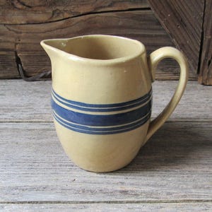 May include: A beige ceramic pitcher featuring a wide horizontal dark blue band around the center, accompanied by two thinner blue stripes above and below the main band. The pitcher has a curved handle and a small pouring spout at the top rim.