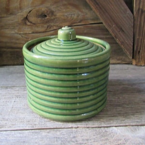 May include: A cylindrical green ceramic jar with a matching lid, featuring a series of horizontal raised ridges that wrap around the entire body and the top of the lid, finished with a smooth, glossy glaze and a small central knob.