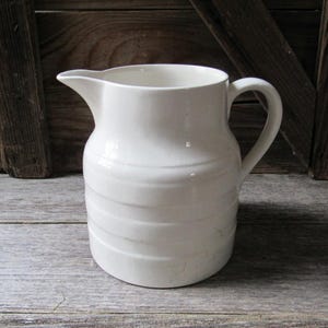 May include: A white ceramic pitcher with a handle and a spout. The pitcher has horizontal ridged detailing around the body. The background is a rustic wooden surface.