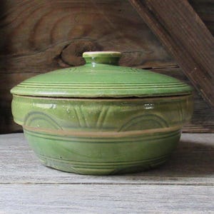 May include: A round, green ceramic bowl with a matching lid. The bowl features a glossy finish and embossed geometric patterns, including horizontal lines and semi-circular arches around the exterior. The lid has a small, centered knob handle.