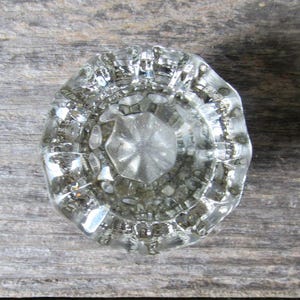 May include: A clear, circular glass door knob featuring a faceted, multi-sided design with a central star-like pattern and a series of concentric, textured ridges radiating outward toward the smooth, rounded outer edge of the hardware piece.