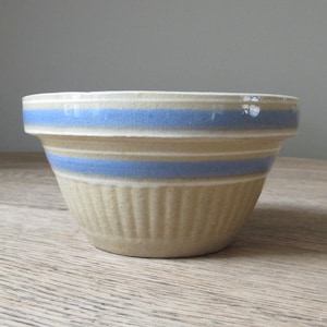 May include: A beige ceramic mixing bowl featuring a wide, flat rim and two horizontal blue stripes encircling the upper portion. The lower half of the bowl displays a vertical ribbed texture, providing a tactile grip and a classic, functional design.