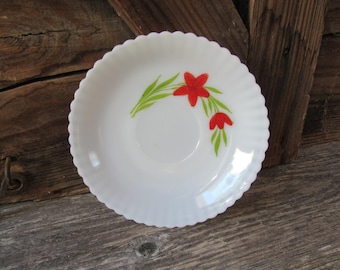 May include: White, scalloped-edge saucer with a floral design. The saucer features a central well and is decorated with red flowers and green leaves. The vintage-style saucer is likely made of ceramic or glass.