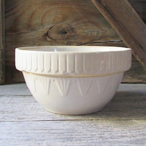 May include: A cream-colored ceramic mixing bowl featuring a wide, reinforced rim with vertical indentations and a lower section decorated with a repeating fan-shaped embossed pattern around the exterior circumference of the vessel.