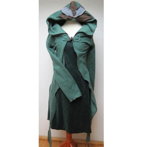 Puede incluir: Una capa con capucha verde con un diseño de capucha en forma de hoja. La capa tiene mangas largas y un dobladillo asimétrico y fluido. La capucha presenta detalles de hojas marrones y grises. La capa se exhibe en un maniquí.