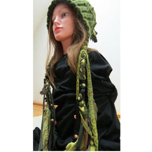 Può includere: Un cappello verde all'uncinetto con lunghe nappe intrecciate. Il cappello è di una tonalità verde vibrante, con un motivo strutturato. Le nappe sono un mix di tessuti verdi e marroni, adornati con piccole perline. Il cappello è un accessorio unico.