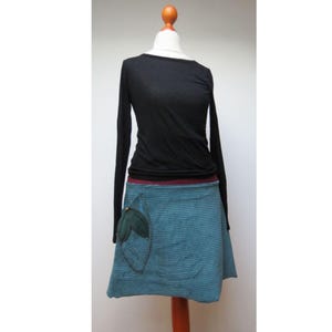 Puede incluir: Una camisa negra de manga larga combinada con una falda a rayas azul turquesa y azul. La falda presenta un bolsillo en forma de hoja verde oscuro. El conjunto se exhibe en un maniquí con un soporte de madera.