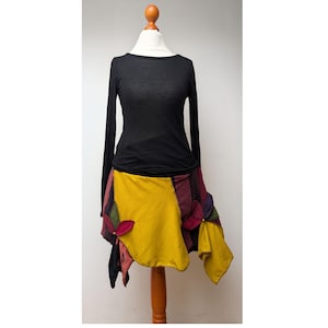 May include: A black long-sleeve top paired with a colorful patchwork skirt. The skirt features yellow, red, and purple fabric panels with decorative flower accents. The outfit is displayed on a mannequin.
