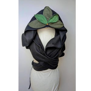 Könnte beinhalten: Schwarzer Kapuzenmantel mit Blättermuster. Die Kapuze hat drei grüne Filzblätter mit einer kleinen Holzperle. Der Umhang wickelt sich um den Körper und wird in der Taille gebunden. Der Umhang ist aus einem weichen, dunklen Material.