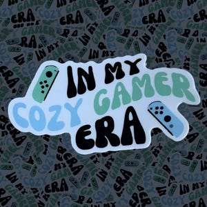 May include: A white sticker with the text "In My Cozy Gamer Era" in black, blue, and green lettering. A blue and green video game controller is depicted on the sticker.