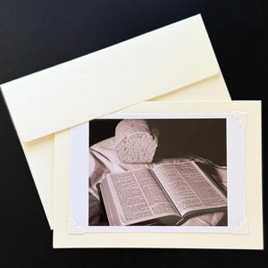 May include: A black and white photograph of a loaf of bread and an open book. The book is lying on a white cloth. The bread is sitting on the cloth behind the book.