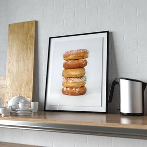 Puede incluir: Impresión enmarcada de una pila de cinco donuts glaseados con diferentes coberturas. Los donuts están en un marco negro sobre un fondo blanco. También se ve un hervidor eléctrico de acero inoxidable y un soporte de madera para utensilios.