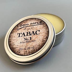May include: A silver tin with a label that reads "LONG RIFLE Soap Co. TABAC No. 1 Artisan Beard Balm 3 fl. oz (89 ml)". The tin is open and the balm inside is a pale yellow color.