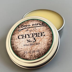 May include: A round metal tin with a green and brown label. The label reads "LONG RIFLE Soap Co. CHYPRE No. 3 Artisan Beard Balm 3 fl. oz. (89 ml)"