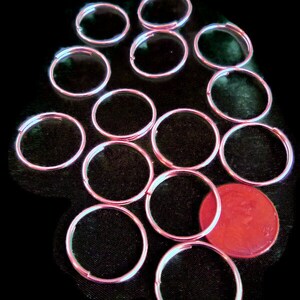 May include: A collection of 13 silver-toned key rings arranged in a circular pattern on a black background. A single red coin is in the center of the circle.