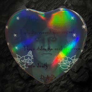 May include: Holographic heart-shaped sticker with a portrait and text. The sticker has a rainbow sheen and features white text and star accents. The text includes phrases like "I have stolen the real crux" and "Face death."