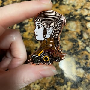 May include: Rose gold enamel pin featuring a profile of a woman with brown hair and a floral design. The woman is wearing a brown and green outfit and has a white face.