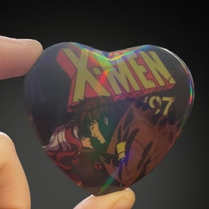 Pode incluir: Botão X-Men '97 em forma de coração com acabamento holográfico. O botão apresenta uma ilustração em quadrinhos de Rogue e Gambit, com o logotipo X-Men em amarelo e vermelho.